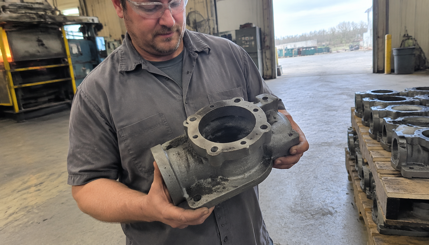 Worker holding a freshly cast gray iron pump housing on a foundry floor